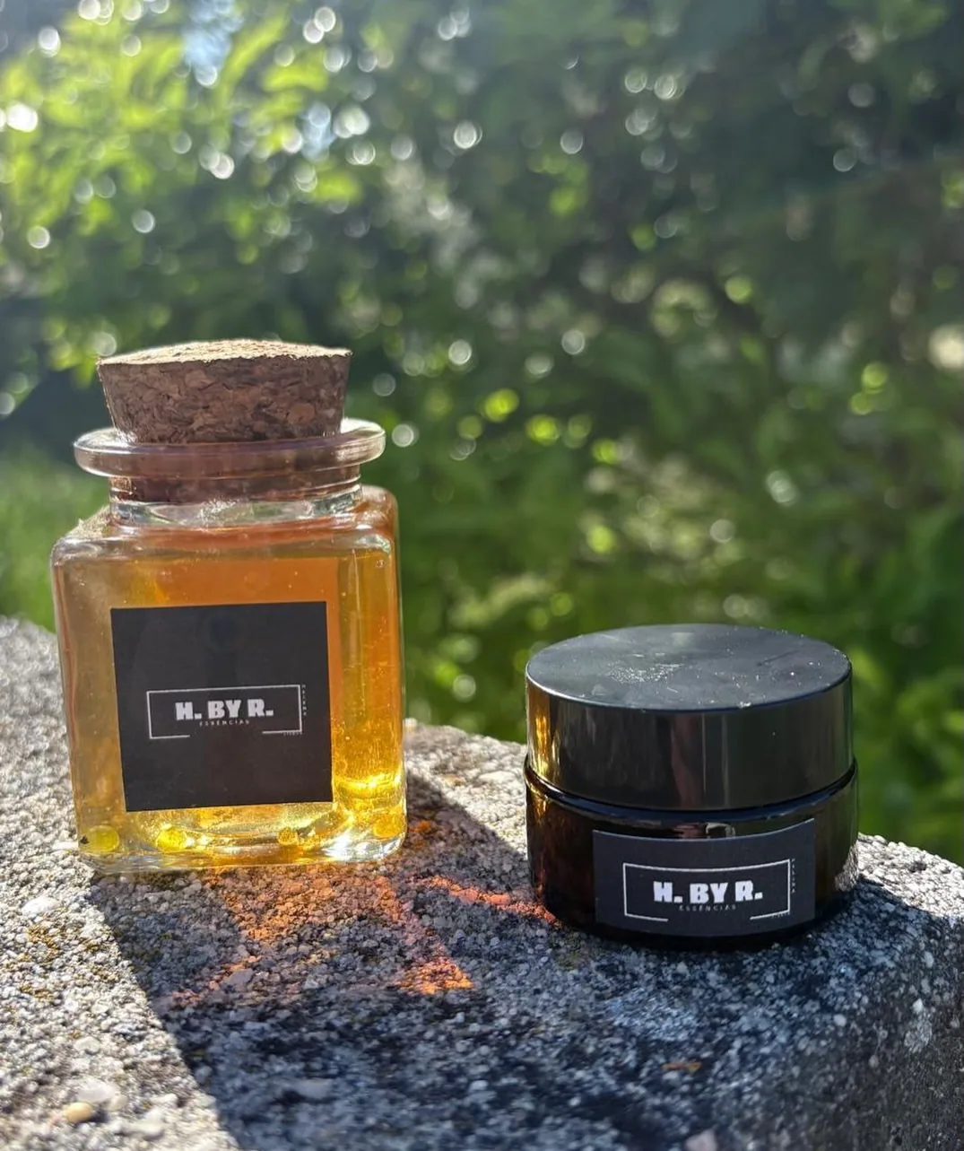 Frasco de essência H.By.R com rolha de cortiça e boião de creme terapêutico, ao sol